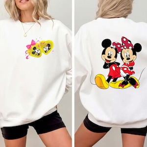 May include: White sweatshirt featuring Mickey and Minnie Mouse. The front displays Mickey and Minnie in a yellow heart with a pink bow. The back shows Mickey and Minnie Mouse in their classic outfits. The sweatshirt is a casual, comfortable style.
