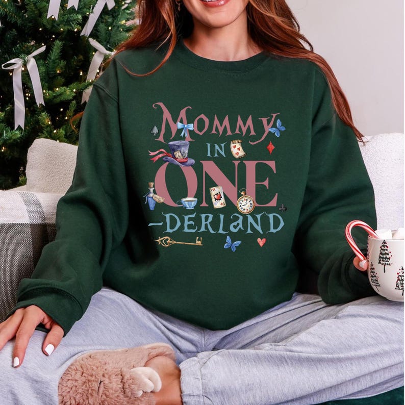 May include: Dark green sweatshirt with the text "Mommy in One -Derland" in a whimsical font. The design includes illustrations of a top hat, playing cards, a pocket watch, and butterflies. The sweatshirt is a comfortable, casual style.