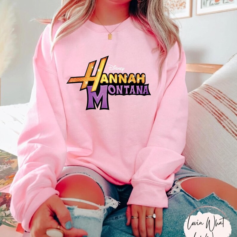 Op de afbeelding: Een lichtroze sweatshirt met het Hannah Montana-logo in geel, paars en zwart. De top met lange mouwen is gemaakt van een zacht materiaal en is geschikt voor vrijetijdskleding. Het Disney-logo staat boven de Hannah Montana-tekst.