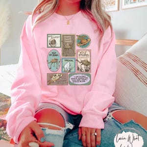 May include: A pale pink sweatshirt adorned with a series of whimsical, retro-style postage stamp graphics. The stamps display diverse illustrations and sayings, such as "Why Stupid?", "Fist My Bump!", and "I am Scary Space Monster".