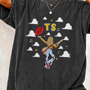 Puede incluir: Una camiseta gris carb&oacute;n de manga corta con un gr&aacute;fico de una persona de pelo largo sosteniendo una guitarra, vistiendo una camisa de cuadros amarillos y pantalones de campana con estampado de vaca. El dise&ntilde;o incluye un sombrero rojo.