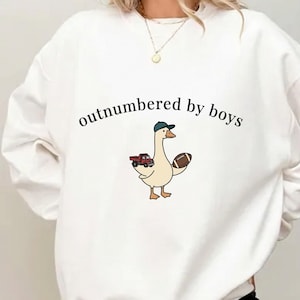 May include: White sweatshirt with the text "outnumbered by boys." A cartoon goose wearing a baseball cap and holding a football and a red truck is printed on the front. The sweatshirt is a casual, comfortable style.