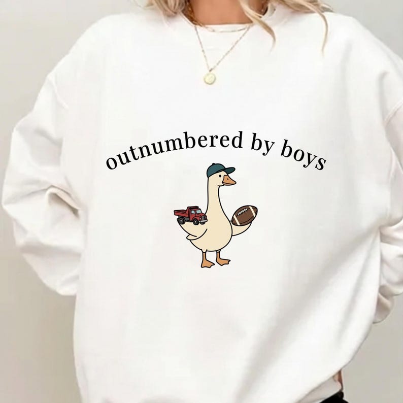 May include: White sweatshirt with the text "outnumbered by boys." A cartoon goose wearing a baseball cap and holding a football and a red truck is printed on the front. The sweatshirt is a casual, comfortable style.