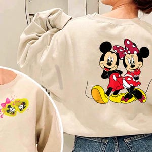 May include: Beige sweatshirt featuring a colorful graphic of Mickey and Minnie Mouse. The cartoon characters are depicted in their classic outfits, Mickey in red shorts and Minnie in a red polka dot dress. The sweatshirt has a crew neck and long sleeves.