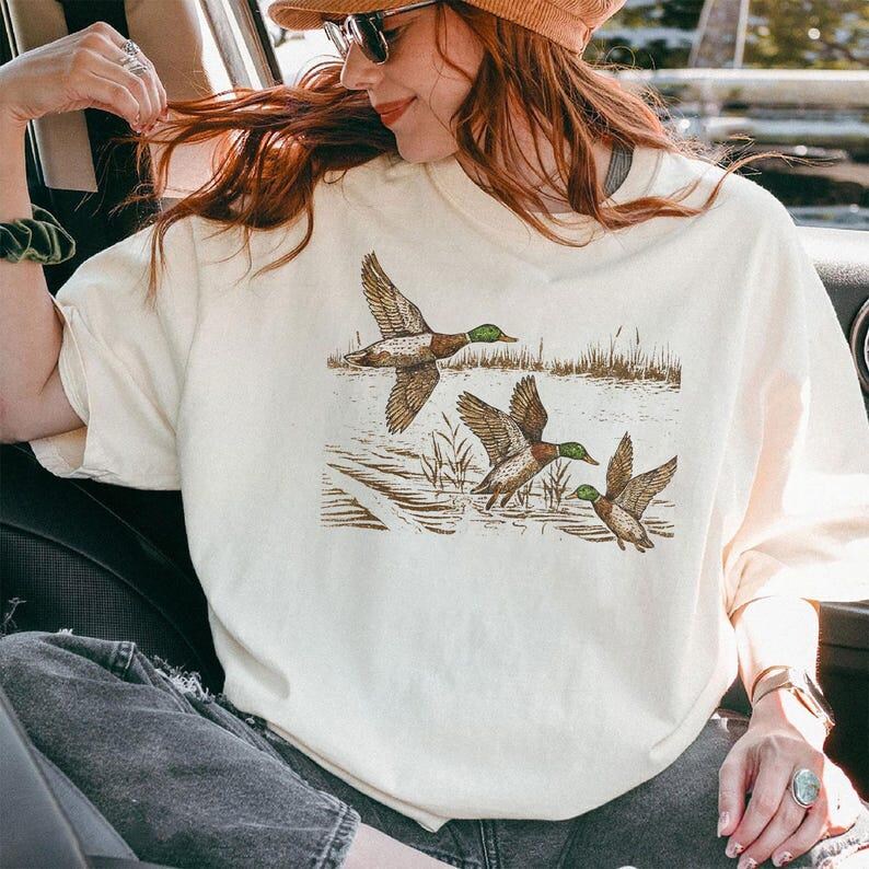May include: Cream-colored sweatshirt featuring a detailed illustration of three ducks in flight over water and reeds. The ducks have brown wings, green heads, and are set against a neutral background. A nature-themed graphic tee.
