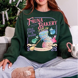 May include: A dark green crewneck sweatshirt featuring a graphic print that reads "Fauna Bakery Since 1959 Once Upon A Dream" with an illustration of a multi-tiered cake, a broom, and three small fairy figures, plus "Baked With Love & Magic".