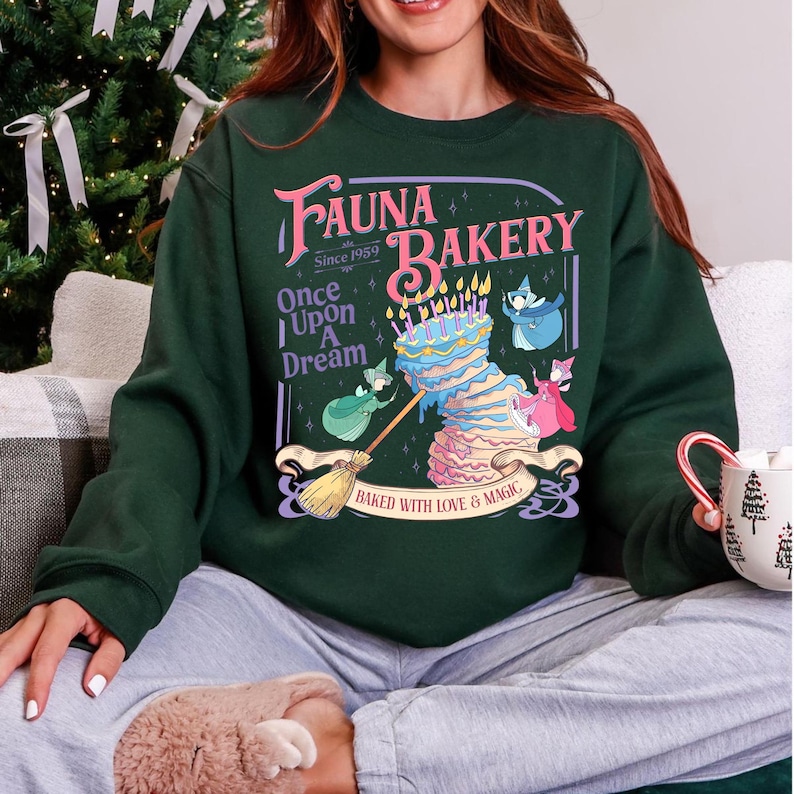 May include: A dark green crewneck sweatshirt featuring a graphic print that reads "Fauna Bakery Since 1959 Once Upon A Dream" with an illustration of a multi-tiered cake, a broom, and three small fairy figures, plus "Baked With Love & Magic".