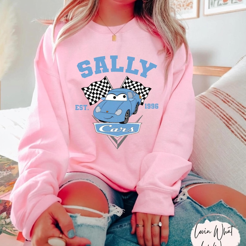 May include: A light pink sweatshirt featuring the name "SALLY" in light blue arched text above a cartoon car graphic. The graphic includes checkered flags and the text "EST. 1996" and "Cars".