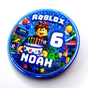 May include: A round blue button with a holographic finish featuring a Roblox character holding a birthday cake with lit candles. The text reads ROBLOX, HAPPY BIRTHDAY, 6, and NOAH. The design includes colourful balloons and confetti elements.
