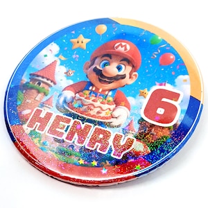 May include: A circular birthday decoration with a cartoon image of Mario holding a cake. The number 6 is prominent, alongside the name "HENRY". The design incorporates a blue sky, stars, balloons, and confetti, creating a festive look.