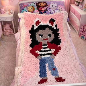 Op de afbeelding: Een roze gehaakte deken met een cartoonfiguur met zwart haar, een rode jas, een blauwe spijkerbroek en rode schoenen. De deken ligt op een wit bed in een roze slaapkamer. Het bed heeft een kussensloop met een patroon.