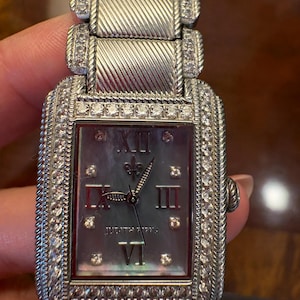 Puede incluir: Un reloj de pulsera rectangular plateado con esfera de nácar. El reloj presenta números romanos como marcadores de hora y está incrustado con pequeñas piedras preciosas transparentes alrededor de la esfera y en la correa. La esfera del reloj muestra la marca "JUDITH RIPKA".