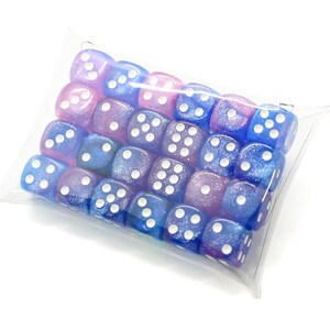 May include: A clear plastic pillow box containing a collection of twenty-four six-sided dice. The dice feature a glittery finish in shades of blue and purple, with white pips indicating the numbers on each of the six faces.