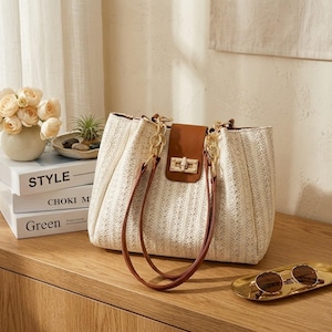May include: A woven beige handbag with brown leather accents and gold-tone chain handles. The bag features a brown flap with a gold clasp. The bag is sitting on a wooden surface, with a pair of sunglasses on a gold tray.