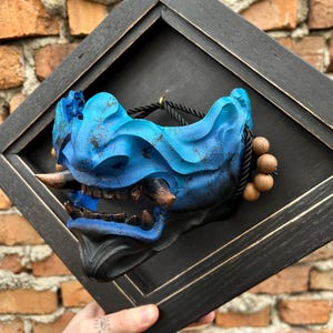 May include: A blue and black Japanese demon mask, framed in a black wooden shadow box. The mask features intricate details, including horns, teeth, and a textured surface. A black rope with wooden beads is attached to the side. The mask is displayed against a brick wall.