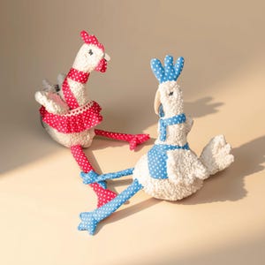 May include: Two plush chicken toys made of white textured fabric. One chicken wears a red polka-dot scarf, skirt, and leg coverings. The other chicken wears a blue polka-dot scarf, vest, and leg coverings. Both have long, dangling legs.
