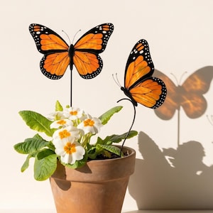 Può includere: Due picchetti da giardino a forma di farfalla monarca arancione e nera sono inseriti in un vaso di terracotta contenente primule bianche con centri gialli. Le farfalle presentano dettagliati motivi alari su sottili aste metalliche.