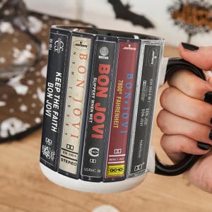 Pode incluir: Caneca de cer&acirc;mica branca com um design de cassetes do Bon Jovi. A caneca exibe t&iacute;tulos de &aacute;lbuns como "Keep the Faith", "Slippery When Wet" e "7800&deg; Fahrenheit". A al&ccedil;a &eacute; preta.
