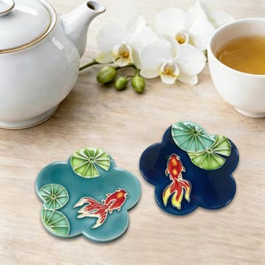 May include: Two ceramic coasters featuring goldfish and lily pad designs. One coaster is teal, the other is navy blue. A white teapot, a cup of tea, and white orchids are in the background.
