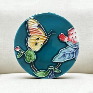 Può includere: Un piatto decorativo rotondo di ceramica color turchese con un design a farfalla e floreale. La farfalla ha ali gialle, arancioni e bianche. I fiori sono rossi e bianchi con foglie blu. Il piatto è probabilmente decorativo.