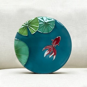 May include: A round, teal-colored decorative plate featuring a red and white goldfish swimming in a pond. Green lily pads are positioned in the upper left. The plate has a glossy finish and is likely ceramic.