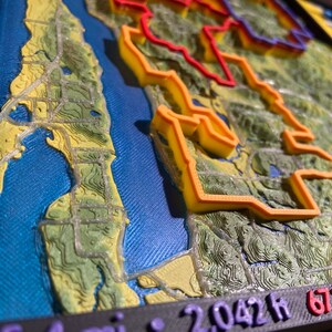 May include: A 3D-printed topographical map with vibrant colors. The map features a blue water area, green and yellow land, and orange and red highlighted areas. The base is black, and the map includes text indicating distance and elevation.