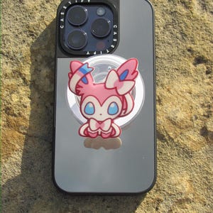 May include: A smartphone case with a black frame and a reflective silver back panel featuring the text CASETiFY around the camera lens. A pink and white cartoon character with blue eyes and ribbon-like appendages is attached to the center of the case.