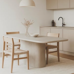 May include: A neutral-toned dining room set featuring a light gray oval table with a rounded base, complemented by four wooden chairs with woven seats. A white pendant light hangs above. A vase of dried flowers and a white bowl sit on the table.