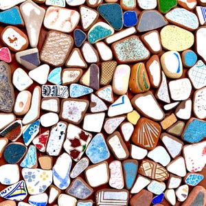 Puede incluir: Un mosaico de piezas cerámicas de forma irregular en varios colores, incluyendo blanco, azul, verde y marrón. Las piezas están dispuestas sobre una superficie marrón rojizo, creando un patrón texturizado y visualmente atractivo. El mosaico es una obra de arte decorativa.