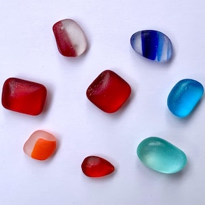 May include: Assortment of sea glass pieces in various shapes and colors, including red, blue, and teal. Some pieces have stripes or color gradients. The sea glass is smooth and rounded, with a frosted appearance.
