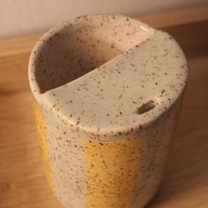 May include: A speckled ceramic toothbrush holder with a yellow stripe. The holder has a unique design with a curved top and a small hole. The neutral color palette and speckled finish give it a rustic, handmade feel.