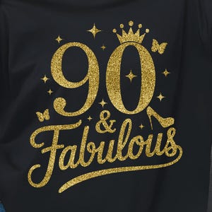 Könnte beinhalten: Schwarzes T-Shirt mit goldenem Glitzerdesign. Das Design zeigt die Zahl "90" mit einer Krone, Schmetterlingen und einem High Heel. Die Worte "& Fabulous" sind in einer Schreibschrift geschrieben.