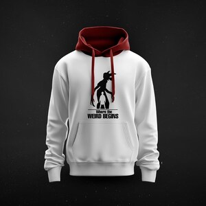 May include: White hoodie with a burgundy hood and drawstrings. The front features a black graphic of a creature and a person with the text "Where the Weird Begins". A comfortable, casual garment.