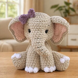 Peut inclure: Éléphant en crochet fait main, beige clair, orné d'un nœud violet. L'éléphant a de grandes oreilles avec un intérieur rose et des détails blancs sur les pieds. Le jouet en peluche est posé sur une surface en bois.
