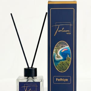 Puede incluir: Un difusor de vidrio transparente con cañas negras y una etiqueta azul marino con texto dorado "Fortum Parfum Fethiye". Una caja azul marino con borde dorado, el mismo texto y una imagen de playa.
