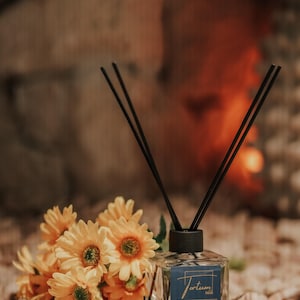 May include: A clear glass diffuser bottle with black reeds and a blue label reading "Tortum Marmaris" sits on small stones. A bouquet of yellow flowers with green centres is next to the bottle. The background is blurred.