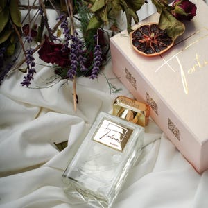 Puede incluir: Un frasco de perfume transparente con tapón y etiqueta dorados, junto a una caja rosa con letras doradas. Flores secas y una rodaja de cítrico seco están dispuestas sobre una tela blanca, creando una estética elegante y natural. El frasco de perfume tiene la palabra "Fortum".