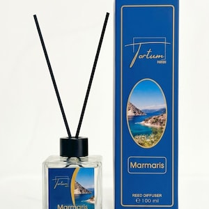 May include: A clear glass reed diffuser with black reeds and a black cap. The diffuser has a label with the text "Tortum Parfum" and "Marmaris" over a scenic image. A blue box with gold trim and the same text and image is next to it.
