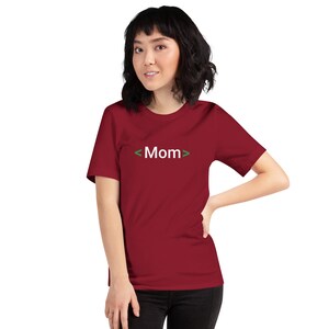 May include: A maroon t-shirt with the word "Mom" in white text, framed by green angle brackets. The shirt is short-sleeved and worn by a person. The person is wearing black pants.