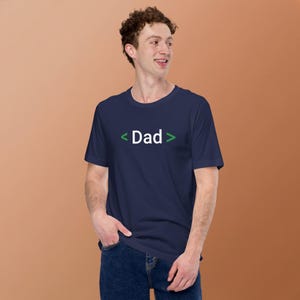 Puede incluir: Una camiseta azul marino con la palabra "Dad" en texto blanco, enmarcada por corchetes verdes. La camiseta es de manga corta y está hecha de un material suave. La persona de la imagen lleva vaqueros azules.