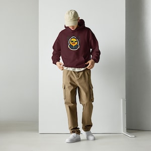 May include: A maroon hoodie with a front pocket and a cartoon duck wearing sunglasses. The duck is outlined in black and yellow. The person is wearing khaki cargo trousers, a beige cap, and white trainers.