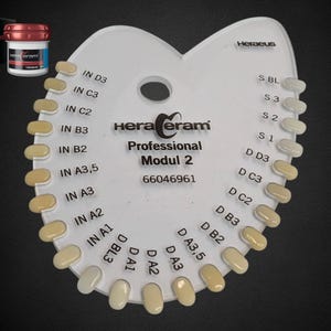 May include: A white plastic dental shade guide with multiple color samples arranged in a semi-circle. The guide is labeled "HeraCeram Professional Modul 2" with various shade codes. A small jar and box with the "HeraCeram" logo are in the upper left corner.