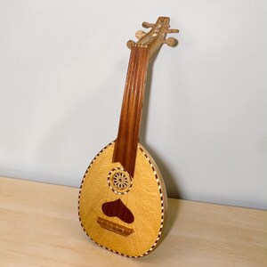 Peut inclure: Un instrument à cordes en bois avec un corps en forme de poire, un long manche et une ouïe circulaire ornée d'une rosace sculptée. Le corps présente un grain de bois clair, une incrustation en forme de cœur et une bordure rayée.