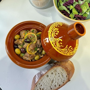 Puede incluir: Un tajine de cerámica marrón con tapa cónica que presenta patrones decorativos amarillos. La base contiene un guiso de carne, aceitunas verdes y negras, y rodajas de limón. Un pan crujiente y un cuenco de ensalada verde fresca se sitúan al lado.
