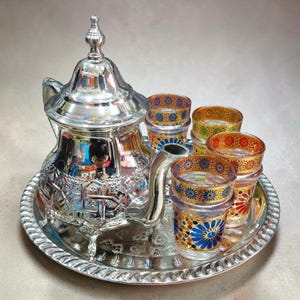 Könnte beinhalten: Ein silbernes Teeservice mit einer dekorativen Teekanne, einem Serviertablett und mehreren kleinen Gläsern. Die Gläser haben farbenfrohe Muster in Blau, Gelb und Rot. Teekanne und Tablett haben verzierte Designs.