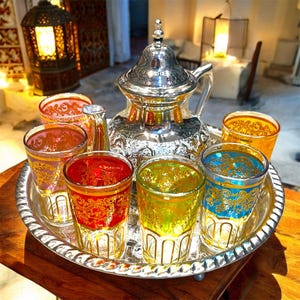 May include: A silver-toned metal teapot with an ornate lid and handle sits on a matching round silver tray, surrounded by five glass tumblers in red, green, blue, orange, and pink, each decorated with intricate gold-coloured filigree patterns.