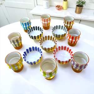 Puede incluir: Una colección de doce recipientes de cerámica, incluyendo varios vasos altos y cuencos más anchos, cada uno con rayas verticales en varios colores como azul, amarillo, rojo, verde, naranja y morado, con interiores blancos y bases de terracota.