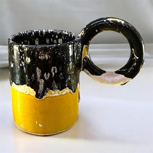 Peut inclure: Une tasse en céramique avec une base jaune vif et une partie supérieure noire brillante ornée de taches blanches. La tasse possède une grande anse circulaire noire qui s'étend sur le côté, créant une silhouette distincte et lisse.