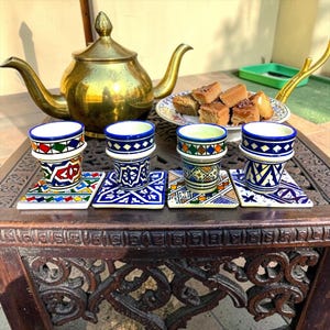 May include: Four ceramic tea cups with intricate blue, green, red, and yellow geometric patterns, each resting on a matching square coaster. A large brass teapot sits behind the cups, and a plate of cubed cake is positioned to the side.