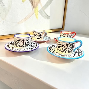 Peut inclure: Un ensemble de quatre tasses à espresso et soucoupes en céramique ornées de calligraphie arabe noire sur fond crème. Chaque ensemble est rehaussé d'un bord coloré distinct, notamment bleu, rouge et gris, formant une collection éclatante.